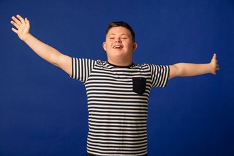 Young person wearing a black and white striped tshirt with arms outstretched and smiling, standing against a plain blue background.