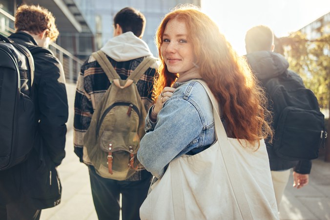 Young girl walking with some boys into a school or college building. She has red hair, wearing a denim jacket and holding a canvas bag. She is looking back and smiling at the camera.