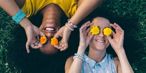 Two young women lying on grass, holding flowers above their eyes and smiling