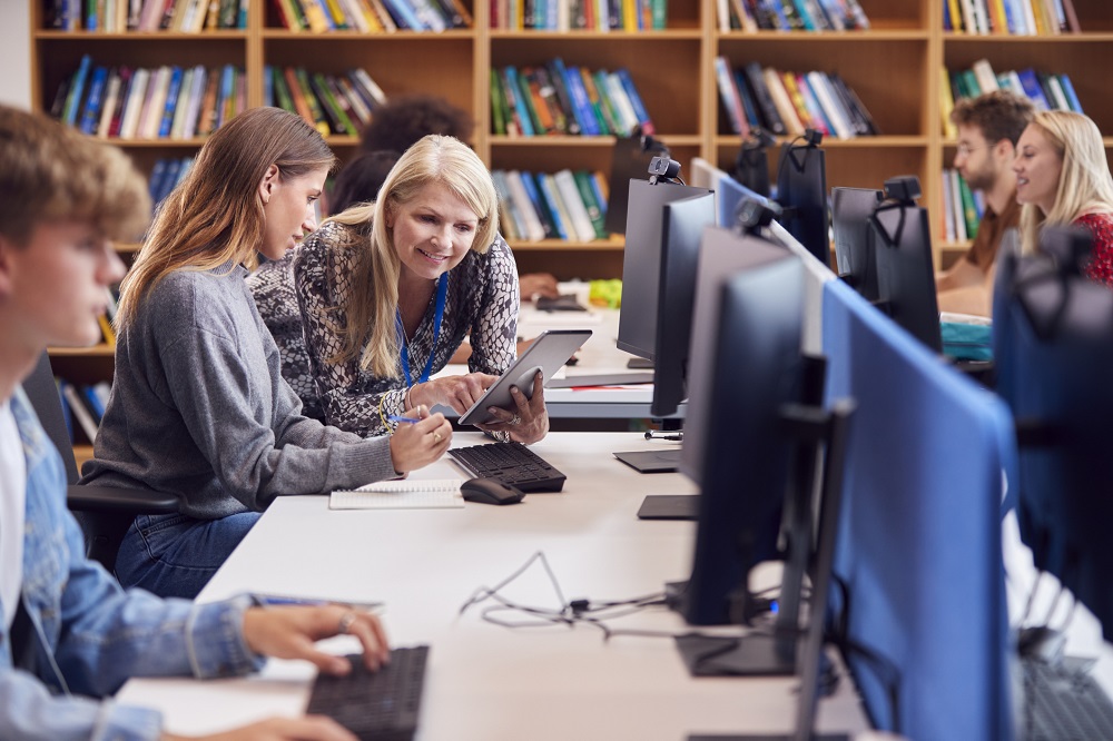 Young person talking to a teacher while looking at a computer in a library..