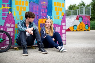 Two young people sitting against a graffitied wall, outside talking.