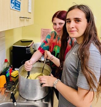 Two young women smiling as they mash potatoes in a large pan in a communal kitchen