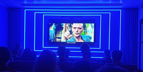 A group of people watch a movie in a dark room with blue neon lighting. The movie shows a character with face paint on a screen.