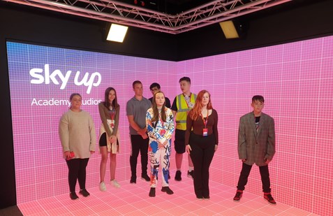 A group of eight young people standing in front of a large screen displaying 'Sky Up Academy Studios.' The background is a pink grid.