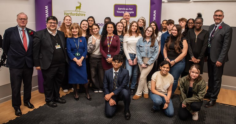 Group of 20 young people of mixed gender and heritage with five adults posing in front of a large stand with Hertfordshire County Council and Services for Young People logos