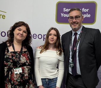 Three people, two female, one male, standing together smiling in front of a stand with a Services for Young People logo