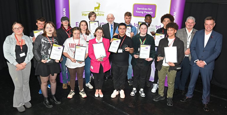 A group of 14 young people of mixed gender and heritage with certificates and three adult males standing in front of a stand with Hertfordshire County Council and Services for Young People logos