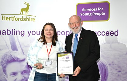 A young woman holding an award being presented with a certificate by an older man standing on front of a stand with Hertfordshire County Council and Services for Young People logos