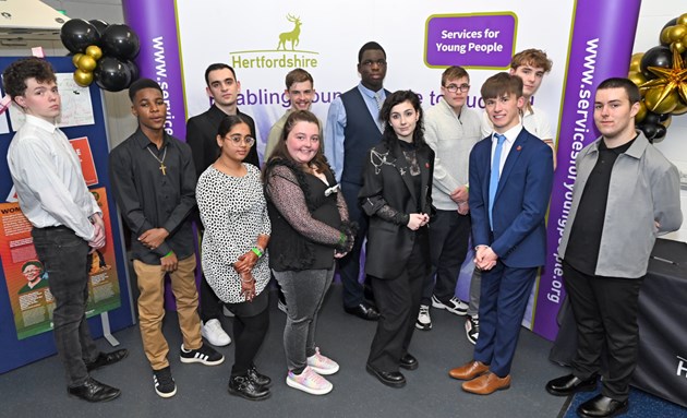 Group of 12 young people of mixed gender and heritage standing on front of a stand with Hertfordshire County Council and Services for Young People logos with black and silver balloons on both sides