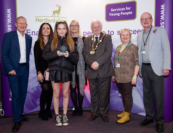 Group of seven people, six adults and one young woman, standing smiling together in front of a stand with Hertfordshire County Council and Services for Young People logos.