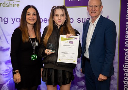 A young woman holding an award and a certificate standing with an adult either side in front of a stand smiling at a celebration event