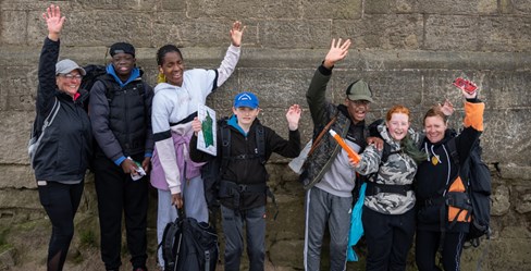 Group of seven people of diverse heritage standing in front of a wall, smiling and waving